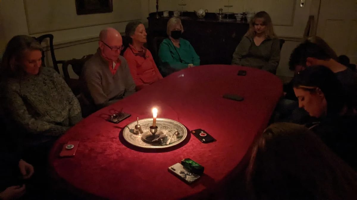 Guests with eyes closed around a red table lit by a single candle and a séance bell during a parlor séance