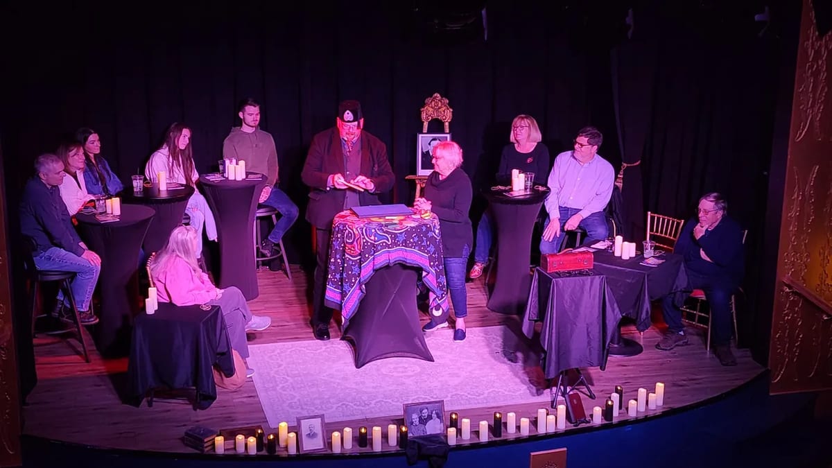 Vince Wilson leading a theatrical séance on a candlelit stage, guests seated around a draped table